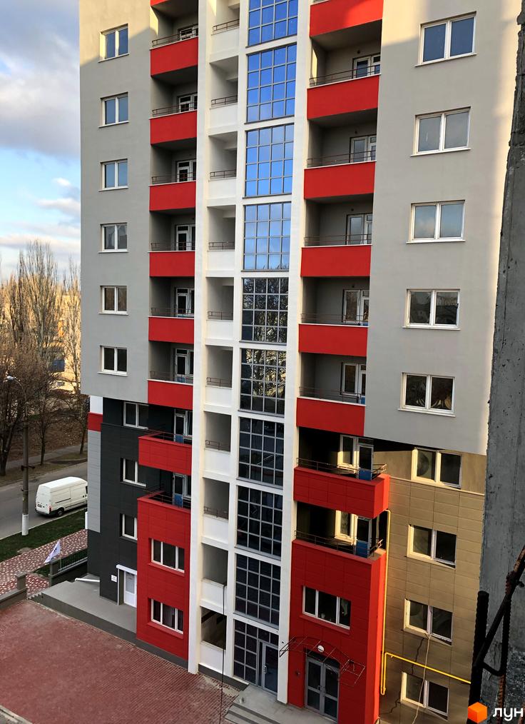 Фасад житлового комплексу Плаза Квартал з червоними та сірими елементами, великими вікнами та балконами. На передньому плані видно бруківку та частину прибудинкової території.