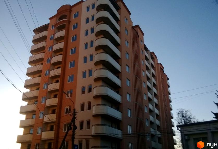 Багатоповерховий будинок з оранжево-бежевим фасадом та численними балконами на тлі блакитного неба. Поруч видно стовп з дротами.
