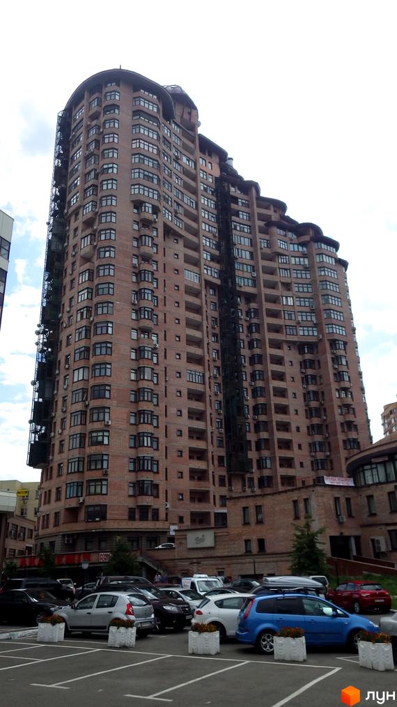 Житловий комплекс на вул. Євгена Коновальця (Щорса), 36в, має багатоповерховий цегляний будинок з балконами. Перед ним видно парковку з автомобілями.