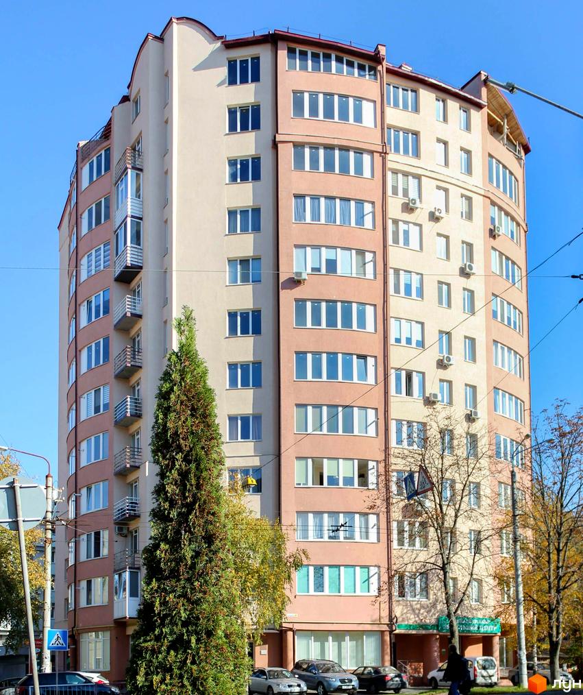 Фото фасаду багатоповерхового будинку за адресою вул. Української Перемоги (Сахарова), 25а. Навколо будинку дерева та припарковані автомобілі.