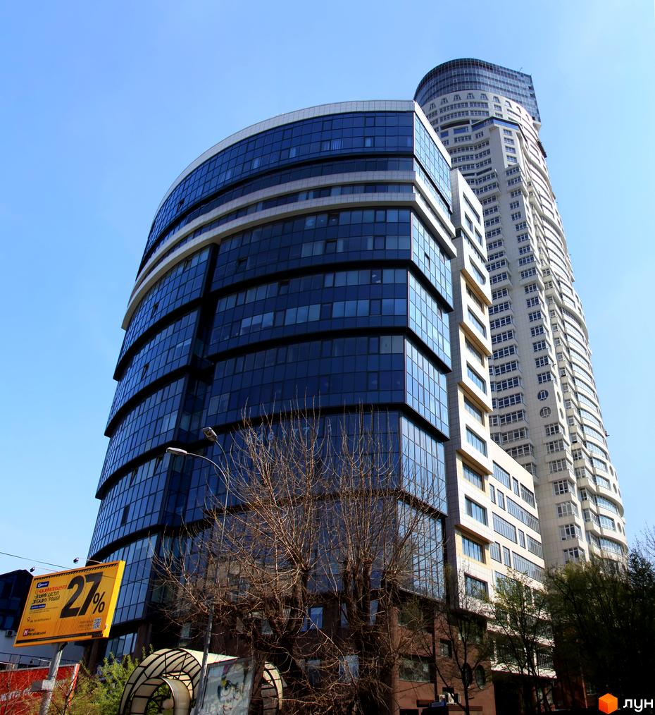 ЖК Carnegie Tower – висока житлова вежа світлого кольору з вигнутим фасадом та численними вікнами. На передньому плані видно дерева та рекламний щит.