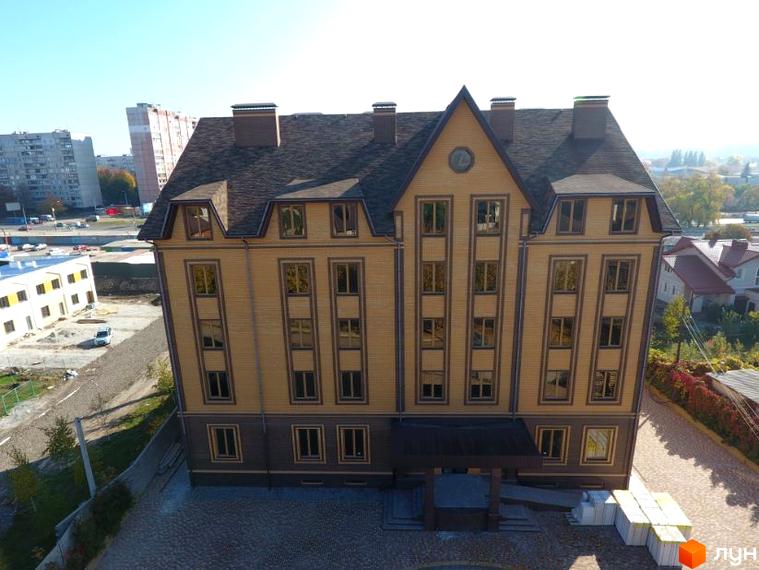 Чотириповерховий будинок Академіка з великими вікнами та коричневим дахом. На прибудинковій території бруківка та пакувальні матеріали.