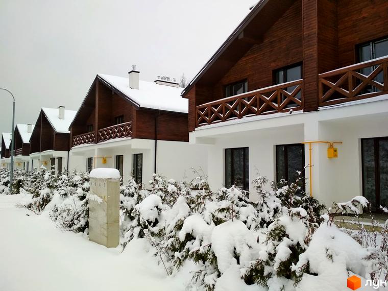 Котеджне містечко КМ Bruhsell у зимовому вбранні. Будинки з дерев'яними фасадами та білими стінами, навколо засніжені ялини та кущі.