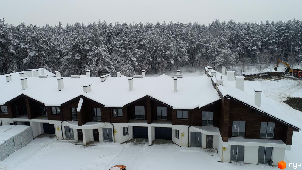 Котеджне містечко Bruhsell взимку: будинки з темно-коричневими фасадами та білими стінами, дахи вкриті снігом. Навколо засніжений ліс та будівельний майданчик з технікою.