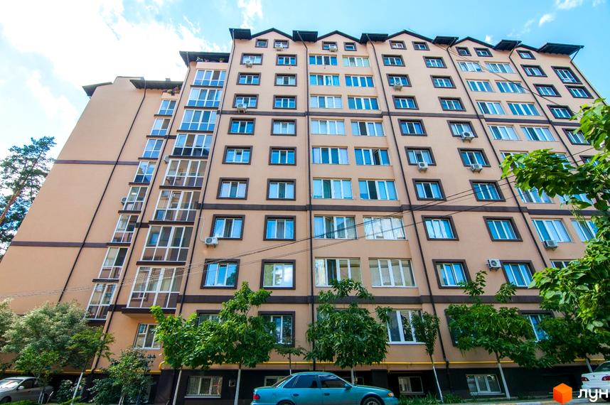 Висотна будівля житлового комплексу з великою кількістю вікон. На першому поверсі видно зелені насадження та припарковані автомобілі.