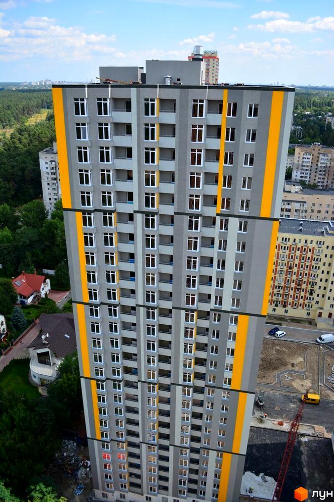 Житловий комплекс Грюнвальд – висотна новобудова сірого кольору з жовтими смугами на фасаді. Навколо будинку – зелені насадження та сусідні будівлі.