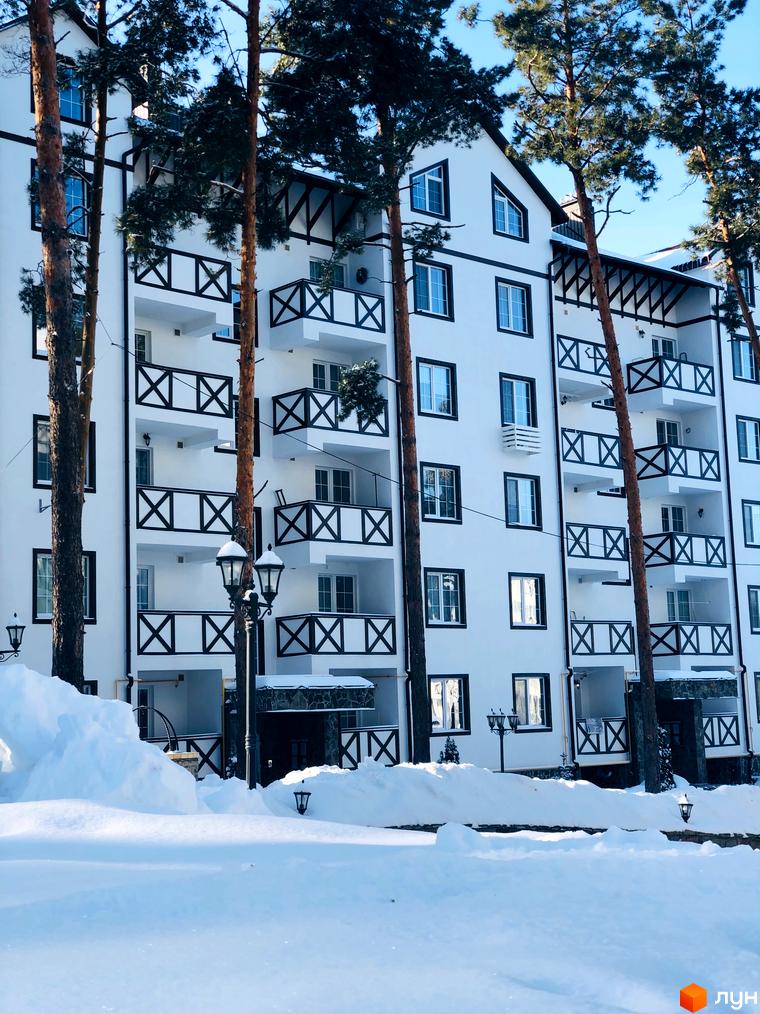 Житловий комплекс DESNA residence взимку. Біла будівля з чорними елементами на балконах та вікнах, оточена соснами. Перед будинком багато снігу та ліхтарні.