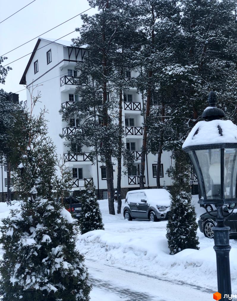 Житловий комплекс DESNA residence у зимовому вбранні. Біла будівля з чорними балконами оточена соснами, вкритими снігом. На території комплексу припарковані автомобілі.
