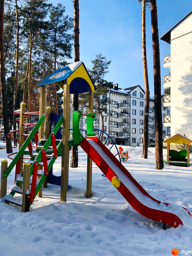 Житловий комплекс DESNA residence з дитячим майданчиком взимку. Сучасна новобудова з балконами та вікнами, оточена соснами.