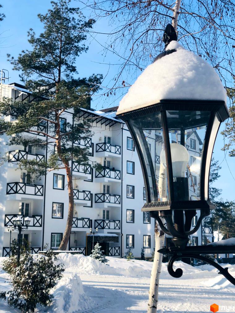 Житловий комплекс DESNA residence взимку. Біла будівля з балконами та дерев'яними елементами на тлі блакитного неба. Навколо засніжена територія з деревами та ліхтарем.