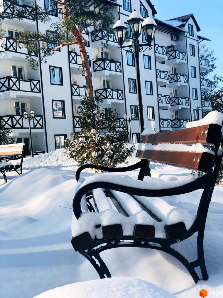 Зимовий вигляд житлового комплексу DESNA residence з білими фасадами та темними балконами. На прибудинковій території розташовані лавки, вкриті снігом, та ліхтарний стовп.
