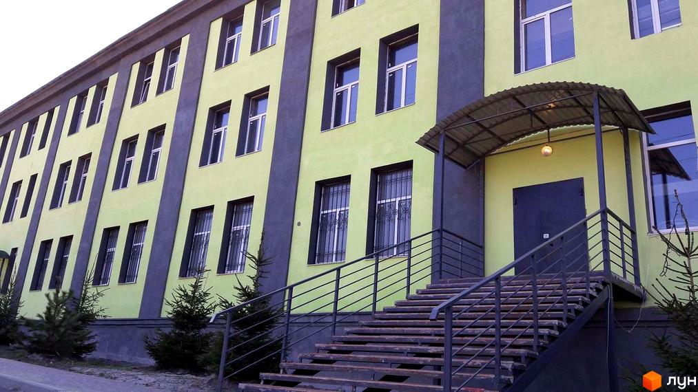 Зелений будинок з великими вікнами та сходами до входу. Перед будинком ростуть ялинки, а над входом навіс.