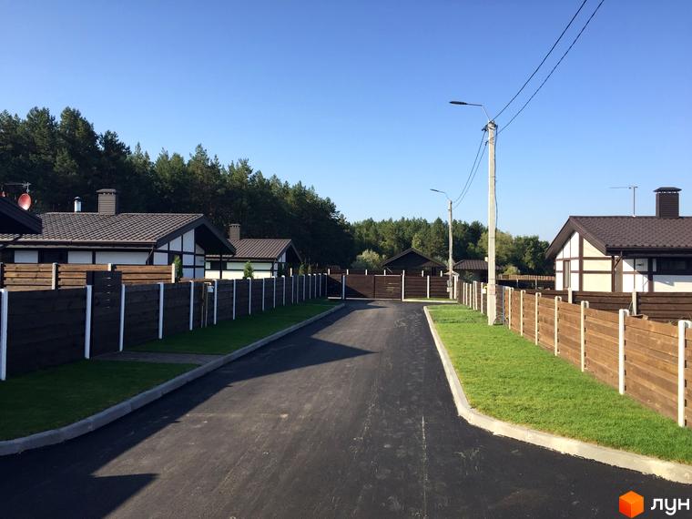 Будинки Котеджного містечка Удачино з темними дахами та світлими фасадами, оточені дерев'яними парканами та зеленими газонами вздовж дороги.