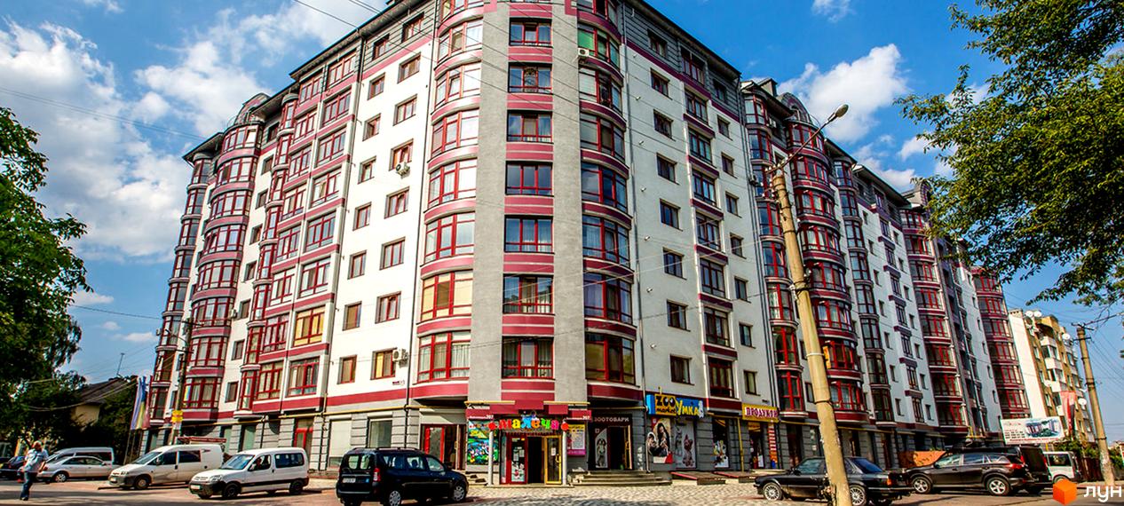 Фото фасаду житлового будинку на вулиці Вовчинецькій та Коломийській. Багатоповерхова будівля з червоними вікнами та магазинами на першому поверсі.