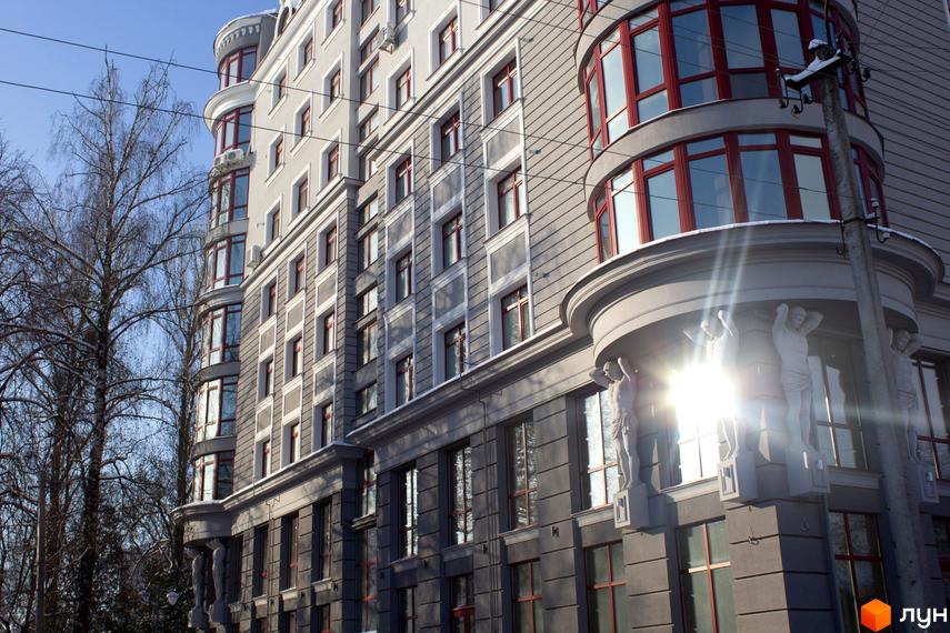 Житловий комплекс Статус: сірий фасад, вікна з червоними рамами та скульптурним декором. Навколо будівлі видно дерева без листя, вкриті снігом.