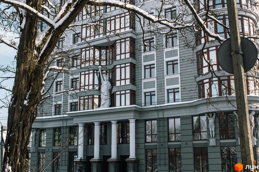 Фасад житлового комплексу Статус із статуєю Свободи та атлантами. Будинок має сірий колір та багато вікон із темними рамами. Навколо будівлі видно гілки дерев, вкриті снігом.