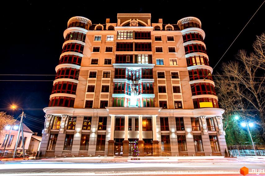 Житловий комплекс Статус - фасадна частина будівлі з виступаючими напівкруглими секціями та великою кількістю вікон. Вхідна група з колонами та підсвіткою. Навколо будівлі видно дерева та вуличне освітлення.