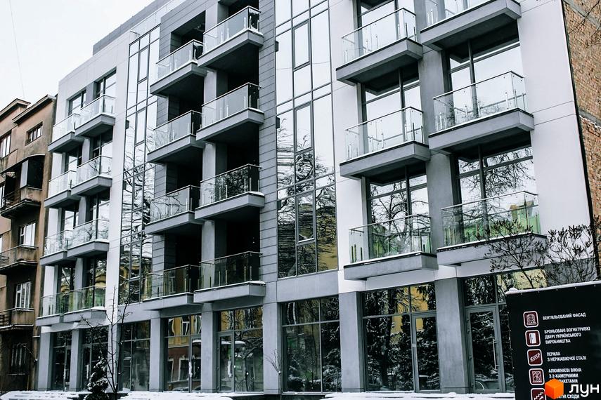 Зовнішній вигляд новобудови ЖК Shevchenko з великими вікнами та скляними балконами. Будинок має сіре оздоблення фасаду та цегляні елементи. Навколо видно дерева та сніг.