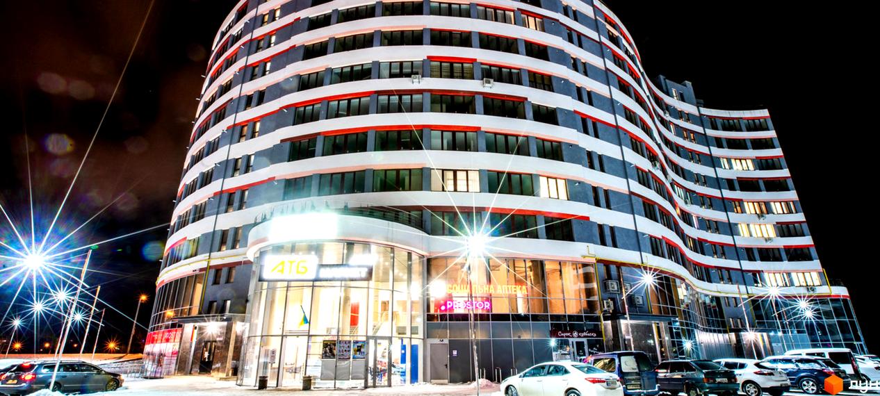 Зображення фасаду багатоповерхового будинку вночі. Житловий комплекс River Park з магазинами та припаркованими авто.