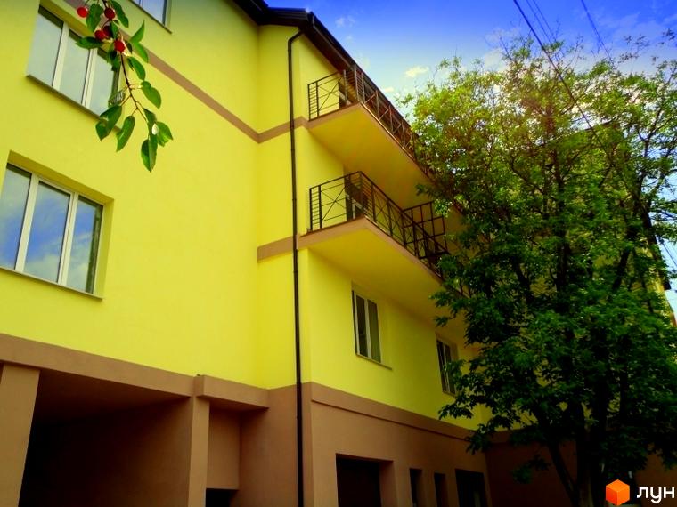 Житловий комплекс Тихорецький: жовта фасадна стіна з коричневими декоративними елементами, балконами та вікнами. Поруч зелені дерева та гілка з ягодами.
