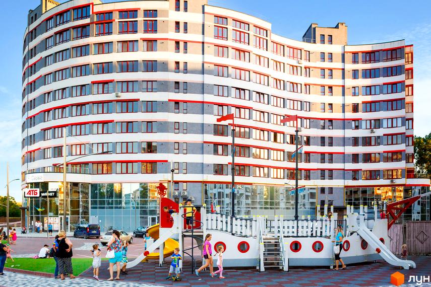 Зображення фасаду Житлового комплексу River Park з дитячим майданчиком у дворі. Багатоповерхова будівля з великою кількістю вікон.