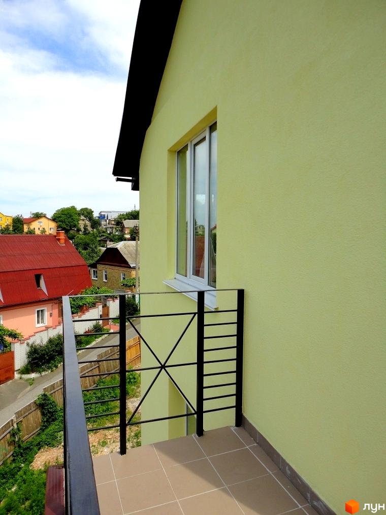 Зовнішній вигляд будинку з жовтим фасадом та вікном, балкон з металевим огородженням. Навколо будинку видно інші житлові будинки та зелень.