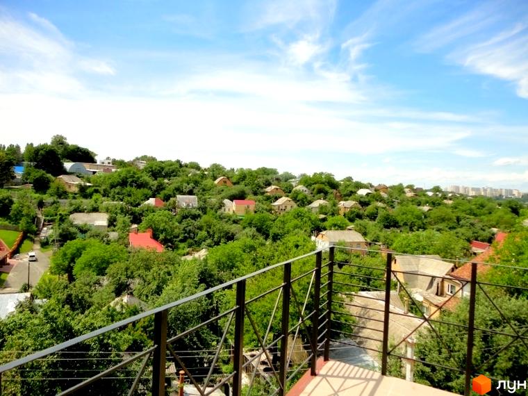 Вид на житловий комплекс Тихорецький з балкона. Зелені дерева та будинки на схилі пагорба під блакитним небом.