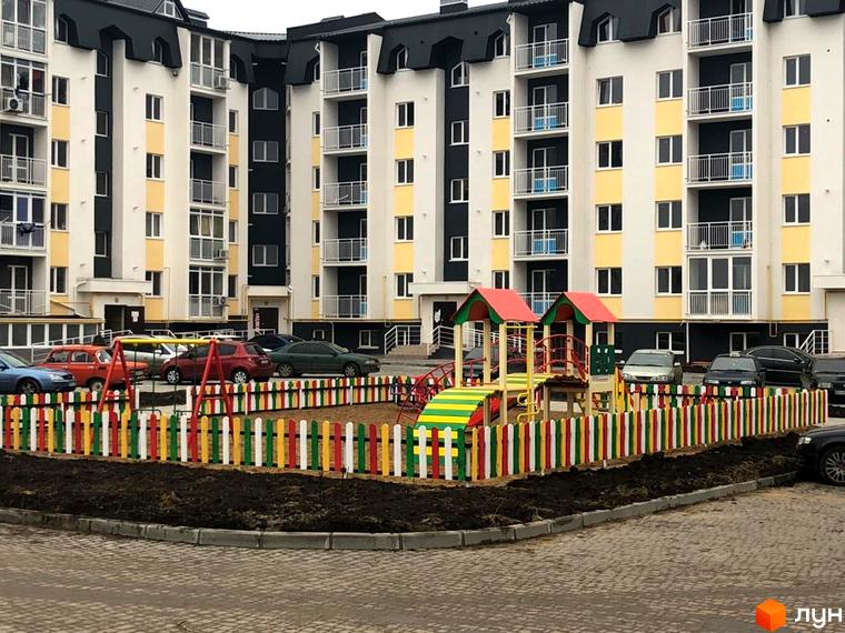 Житловий комплекс Новосел: багатоповерховий будинок з білим, чорним та жовтим фасадом, балконами. Перед ним дитячий майданчик з різнокольоровою огорожею.