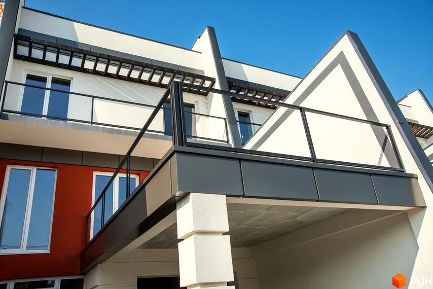 Фасад таунхаусів Капітель: білі стіни, темно-сірі та червоно-коричневі вставки, балкони зі скляними перилами та дерев'яними елементами.