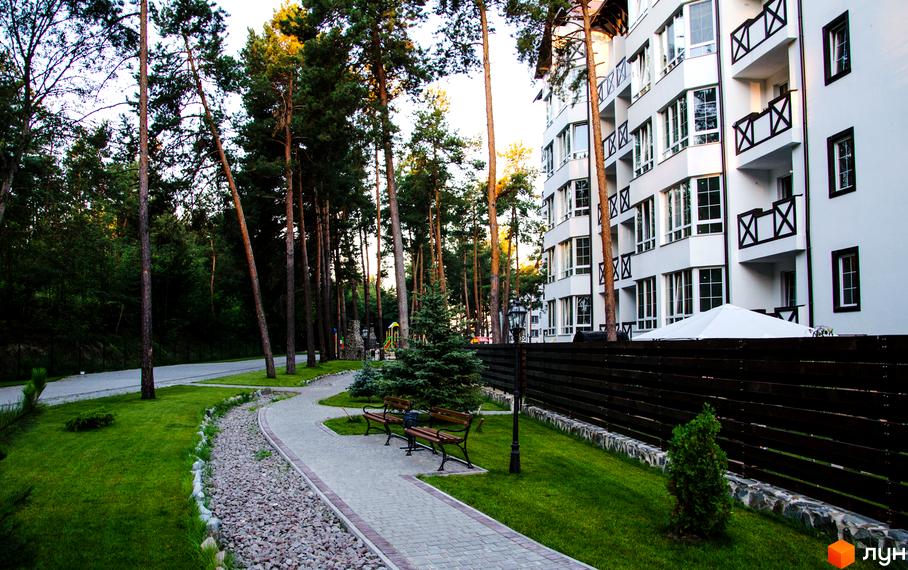 Житловий комплекс DESNA residence з білою фасадною частиною та балконами з дерев'яними елементами. Навколо комплексу розташовані зелені насадження та доглянута прибудинкова територія з доріжками та лавками.