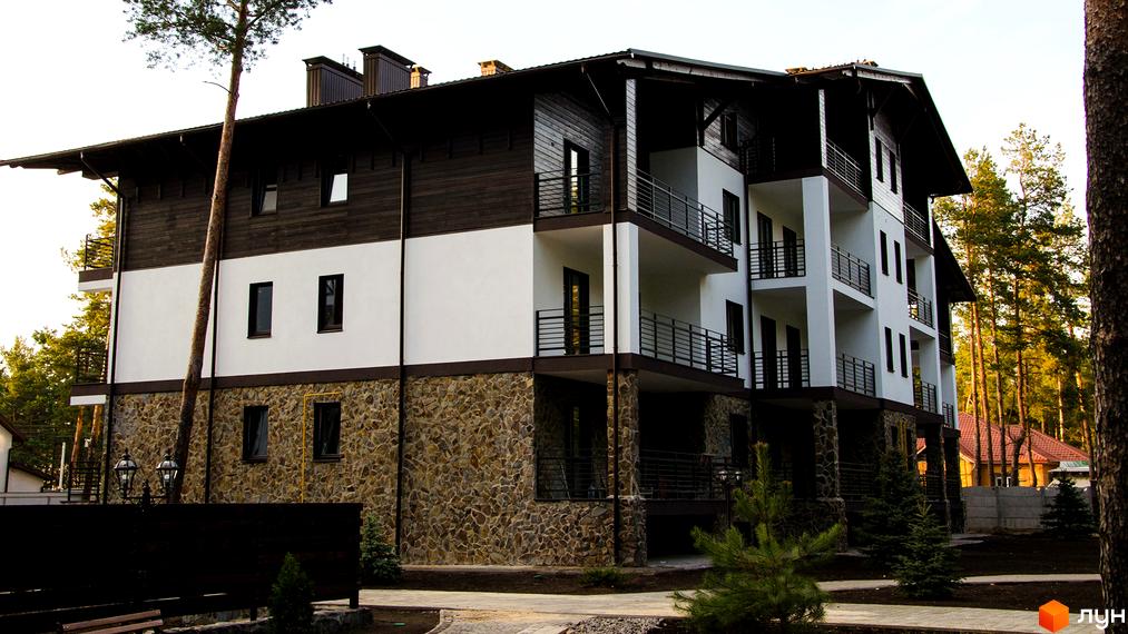 Житловий комплекс DESNA residence - триповерхова будівля з білими стінами, темними дерев'яними елементами та кам'яним цоколем. Навколо будівлі розташовані дерева та зелені насадження.