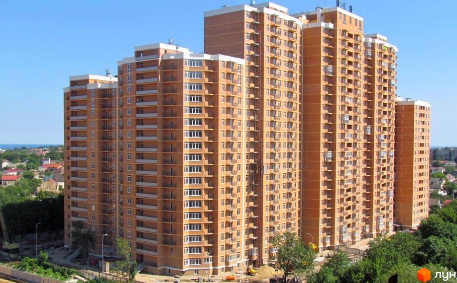 Зовнішній вигляд житлового комплексу Дмитріївський, що складається з висотних будинків з цегляним фасадом та балконами. Навколо комплексу видно зелені насадження та елементи будівельного майданчика.