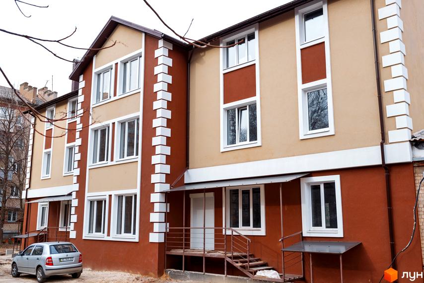 Зовнішній вигляд триповерхового будинку ЖК Дім на Набережній з коричневими та бежевими фасадами, білими вікнами та декоративними елементами. Біля будинку стоїть сріблястий автомобіль.