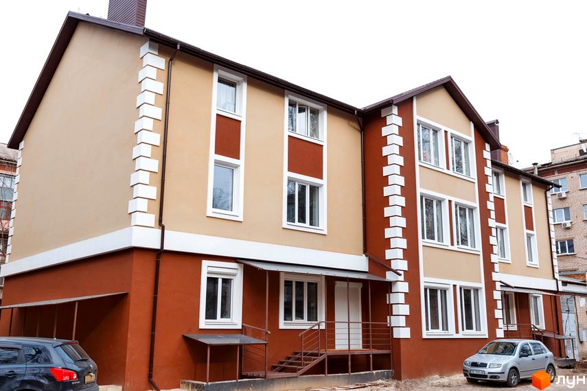 Зовнішній вигляд новобудови ЖК Дім на Набережній з коричневим та бежевим фасадом, білими декоративними елементами та вікнами. Поруч припаркований сріблястий автомобіль.