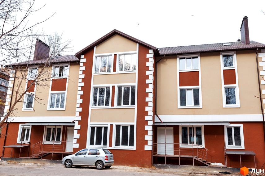 Фасад будинку ЖК Дім на Набережній з коричневими та бежевими елементами, білими вікнами та декоративними елементами. Поруч припаркований сріблястий автомобіль.