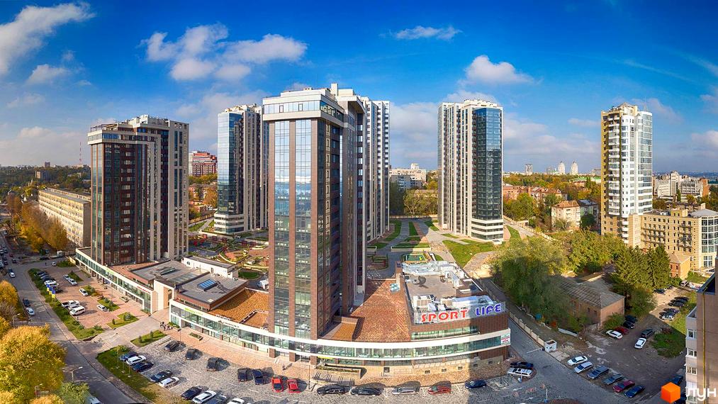 Високі будинки ЖК Панорама з скляними фасадами на тлі блакитного неба. На території комплексу є парковка та зелені зони для відпочинку.