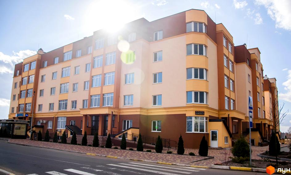 Житловий комплекс Дмитрівський - багатоповерхова новобудова з жовто-бежевим фасадом та коричневими елементами. Перед будинком розташована доглянута прибудинкова територія з деревами та пішохідною доріжкою.