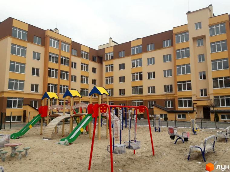 Новобудова ЖК Дмитрівський з дитячим майданчиком. Багатоповерховий будинок світло-жовтого та коричневого кольору з великими вікнами.