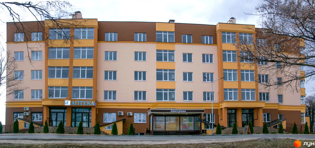 Новобудова ЖК Дмитрівський з фасадом у жовто-бежевих тонах та коричневою обробкою верхніх поверхів. Поруч розташована зупинка громадського транспорту та зелені насадження.