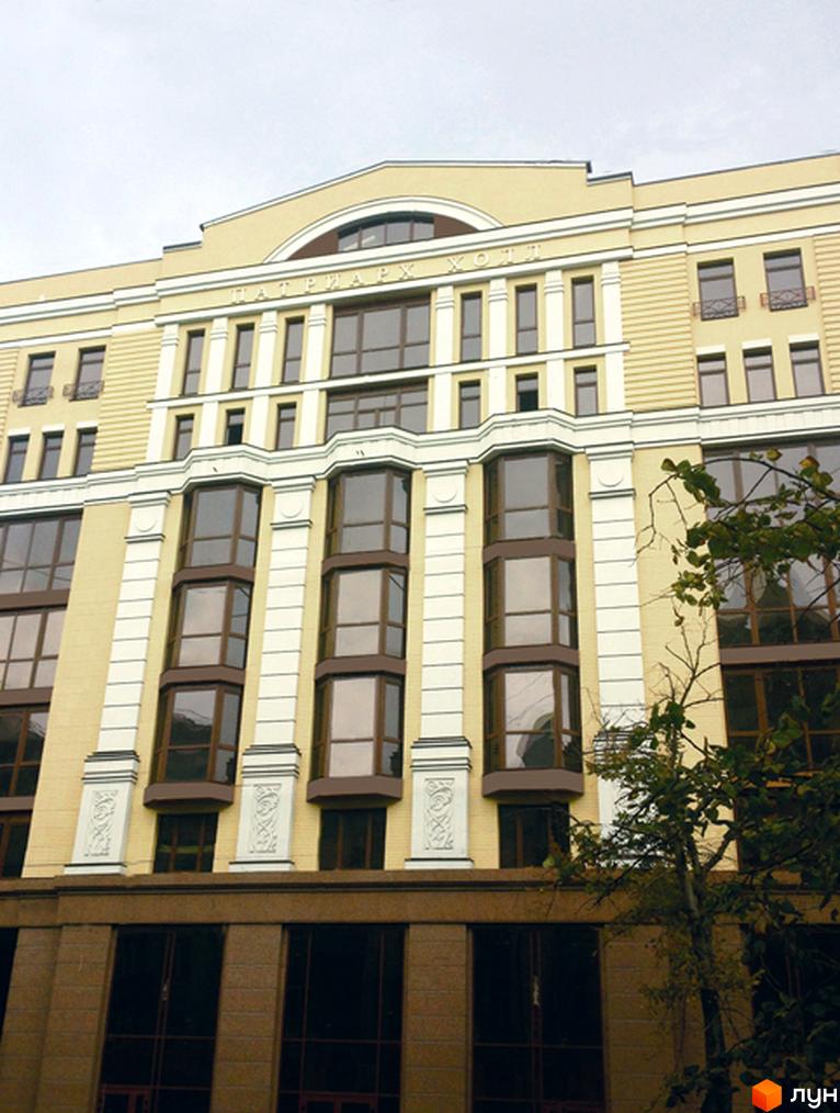 Зображення фасаду Клубний будинок „Патріарх Холл“. Висока будівля з великими вікнами та світлим фасадом. Перед будівлею росте зелене дерево.