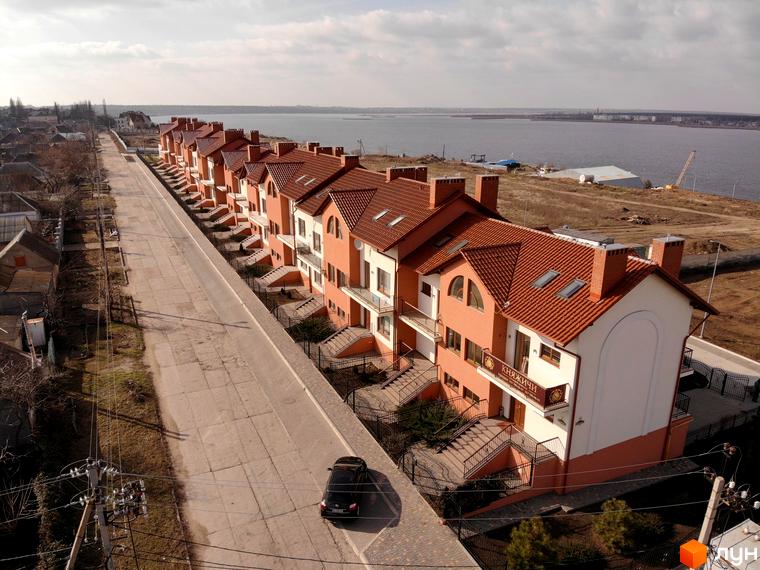 Ряд будинків з червоною черепицею та цегляними фасадами в Котеджному містечку Княжичі, з видом на водойму. На вулиці стоїть чорний автомобіль.