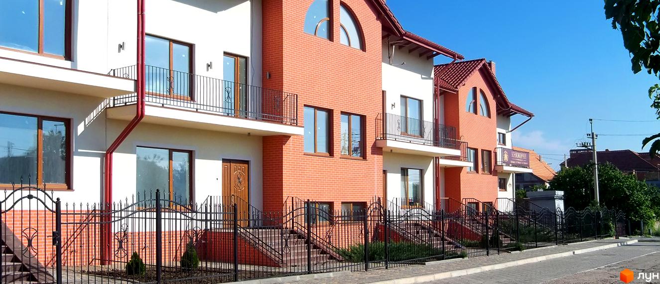 Зображення фасаду будинку в Котеджному містечку Княжичі з цегляними стінами, балконами та металевою огорожею. На прибудинковій території висаджені кущі.