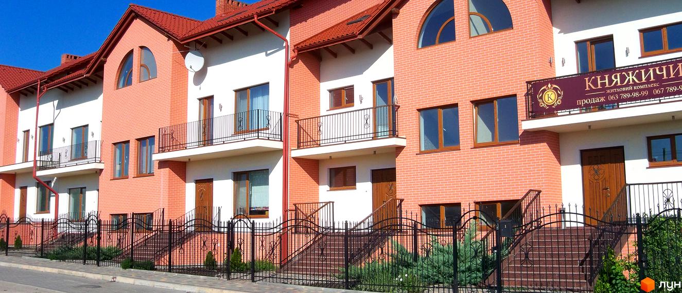 Зовнішній вигляд котеджного містечка Княжичі з цегляним фасадом та червоним дахом. Огороджена прибудинкова територія з доглянутими рослинами.