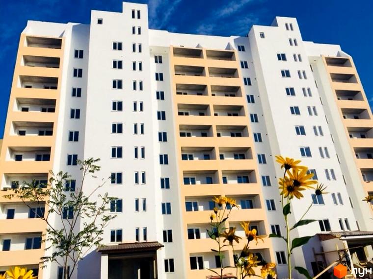 Висотна будівля ЖК Едельвейс з балконами та великою кількістю вікон на фоні блакитного неба. На передньому плані квіти та зелень.
