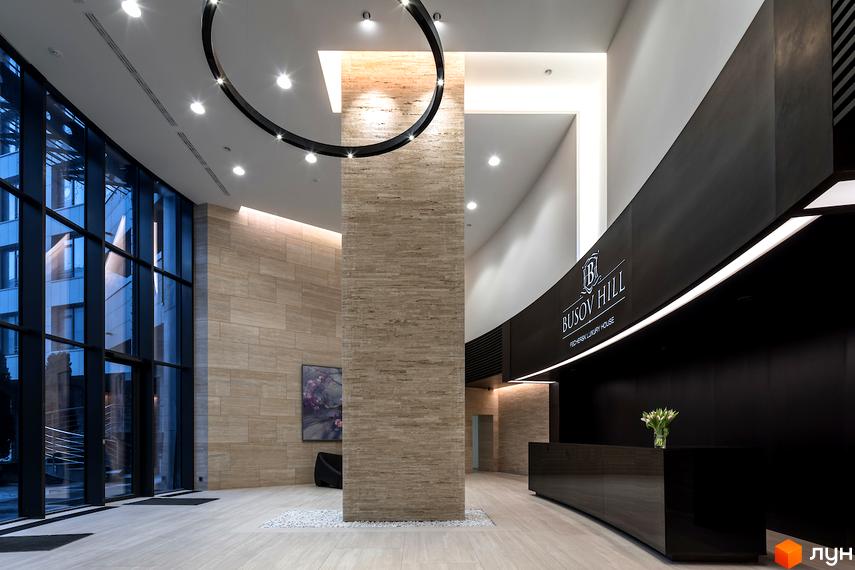 Дизайн інтер'єру холу житлового комплексу Busov Hill. Просторий вестибюль з великими вікнами та центральною колоною, оздобленою натуральним каменем. Стіни оздоблені світлим мармуром, підлога – дерев'яною плиткою.