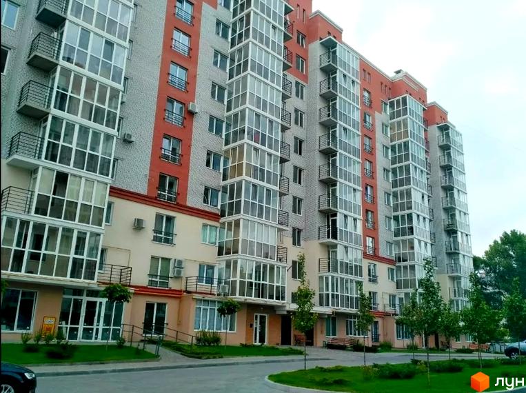 Висотна будівля житлового комплексу з скляними балконами та червоними вставками на фасаді. На прибудинковій території зелені насадження та асфальтована дорога.