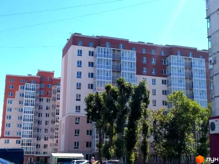 Зовнішній вигляд новобудови на пров. Героїв Бресту, 75а, з фасадами світло-бежевого та темно-червоного кольорів, скляними балконами та зеленими деревами на передньому плані.