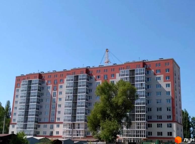 Багатоповерховий будинок з червоним дахом та скляними балконами. На території видно дерева та блакитне небо.