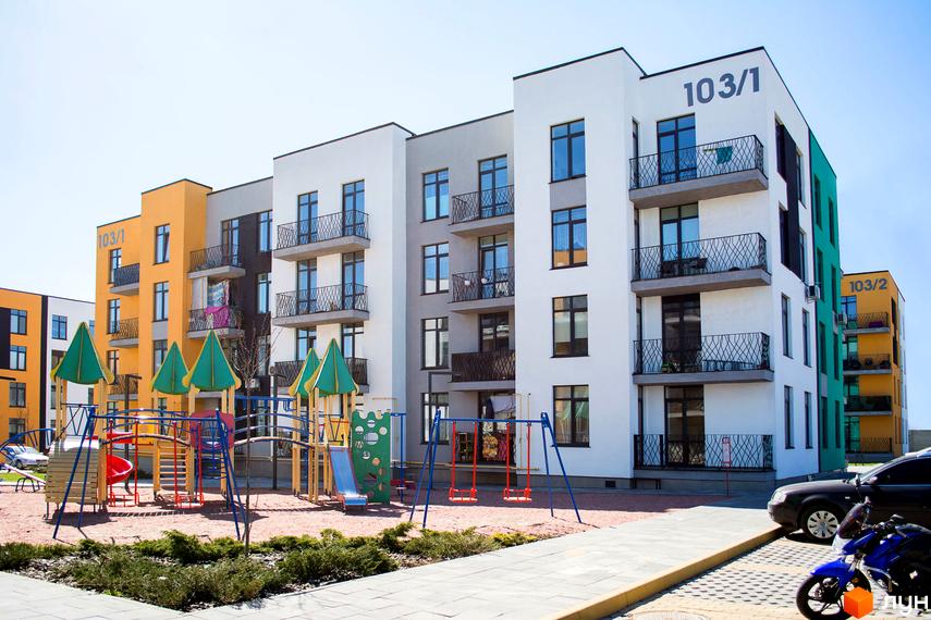 Житловий комплекс Європейка: фасади будинків у білому, жовтому та сірому кольорах, з балконами та вікнами. Поруч дитячий майданчик з гірками та гойдалками.