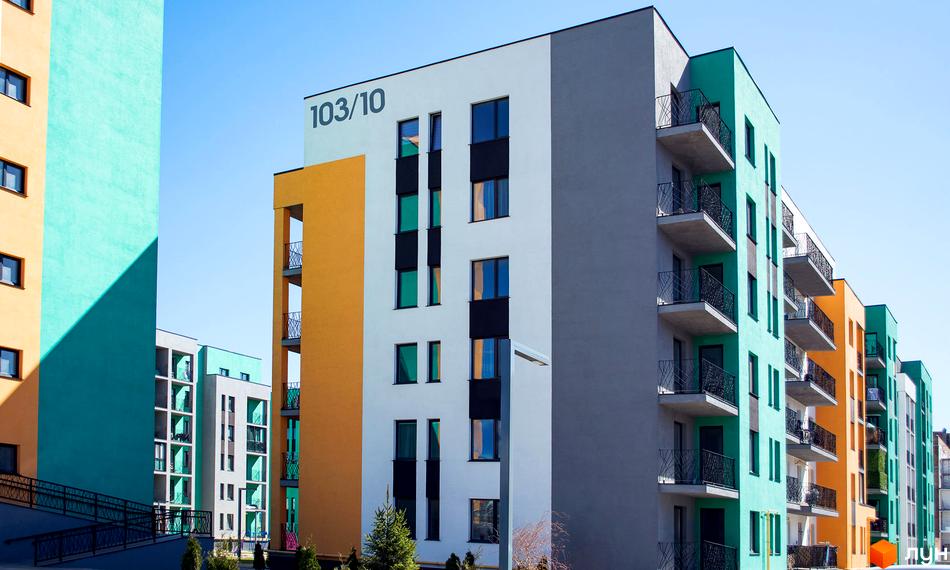 Фасад житлового комплексу Європейка з яскравими кольорами: білий, сірий, жовтий та бірюзовий. Будинки мають балкони та великі вікна. Навколо видно зелені насадження та елементи благоустрою.